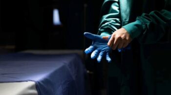 Conheça mais detalhes sobre o funcionamento do IML! Close-up das mãos de um profissional de saúde vestindo luva cirúrgica azul em sala de exames do funcionamento do IML.